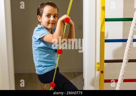 Das Mädchen lächelt, spielt und sportet auf der Treppe und der schwedischen Mauer im Kinderzentrum in speziellen engen Klamotten und führt eine gesunde Stockfoto