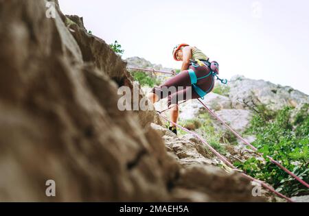 Aktive Klettererin mit Schutzhelm Abseilen von der Felswand mit Seil mit Sicherungsvorrichtung und Klettergurt. Aktive Extremsportzeit Stockfoto