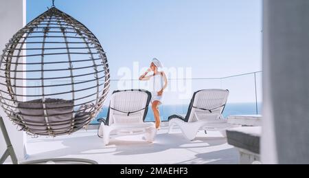 Nach der Dusche schöne Frau mit Haaren und Körper Handtuch wickeln stehen auf offener Terrasse mit Sonnenliegen, Hängesessel genießen Seeslandschaft und blau sauber Stockfoto