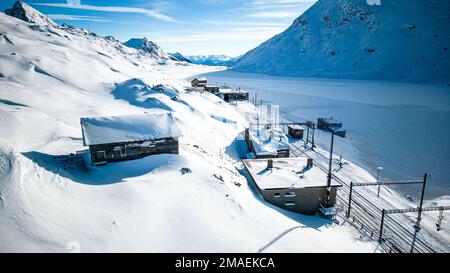 Blick aus der Vogelperspektive auf Holzhütte und Bahnhof sowie im Winter mit Schnee bedeckte Strecken, umgeben von Bergen im alpinen Pass in der Schweiz. Stockfoto