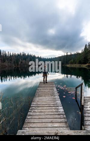 Ein weißer junger Mann mit einer Mütze, die eine Kamera hält, steht auf einem Pier in der Nähe eines Sees mit Waldhintergrund. Stockfoto