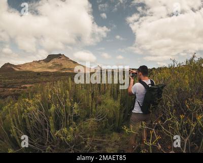 Kaukasischer junger Mann mit dunklen Haaren und Tasche fotografiert Landschaft mit Kakteen und Bergen in Arona, Teneriffa. Stockfoto