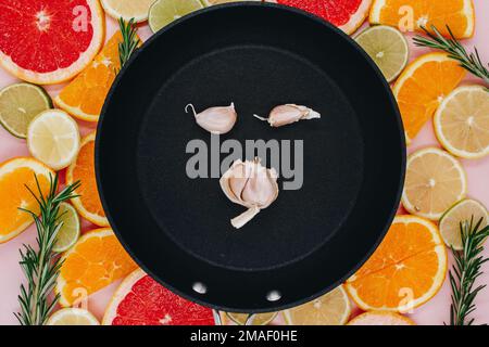 Schwarze Bratpfanne mit Knoblauch auf Zitruskräutern. Orangen, Grapefruit, Zitrone, Limette auf rosa Hintergrund. Flach verlegt Stockfoto