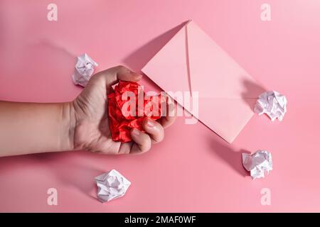 Die Hand eines aggressiven und frustrierten Mannes, der das Herz aus zerknittertem Papier presst. Briefumschlag mit verstreuten Papierstückchen. Brief an einen geliebten Menschen über Valentin Stockfoto