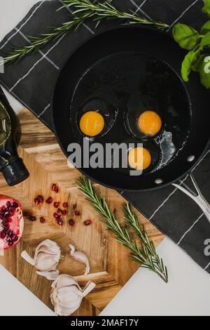 Schwarze Pfanne auf einem schwarzen Handtuch mit drei rohen Eiern neben einem Holzbrett mit rotem Granatapfel. Stockfoto