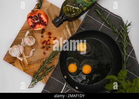 Schwarze Pfanne auf einem schwarzen Handtuch mit drei rohen Eiern neben einem Holzbrett mit rotem Granatapfel. Stockfoto