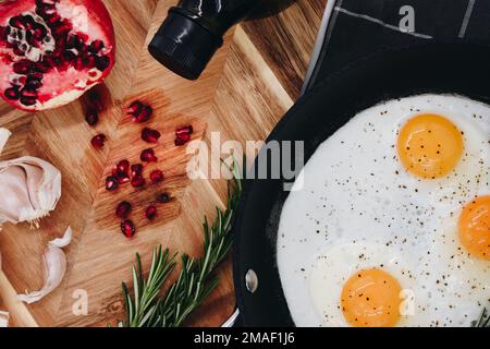 Schwarze Pfanne auf einem schwarzen Handtuch mit drei gebratenen Eigelb neben einem Holzbrett mit rotem Granatapfel. Stockfoto