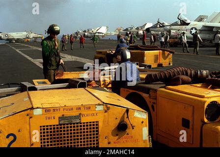 Drei Besatzungsmitglieder stehen mit zwei Schleppern MD-3 (rechts) und einem mobilen Elektrokraftwerk NC-2 (links) während des Cockpit-Betriebs an Bord des Flugzeugträgers USS CORAL SEA (CV-43) bereit. Im Hintergrund befinden sich ein F-4 Phantom II Flugzeug von Marine Fighter Attack Squadron 531 (VMFA-531) (links) und A-7 Corsair II und A-6 Intruder auf der rechten Seite. Land: Unbekannt Stockfoto