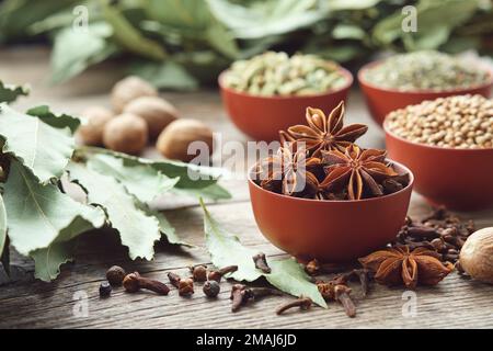 Eine Schüssel Anissterne. Schüsseln mit aromatischen Gewürzen - Koriander, Kardamomschoten im Hintergrund. Handschuhe, Lorbeerblätter, Muskatnuss auf dem Tisch. Inhaltsstoffe für die Gesundheit Stockfoto