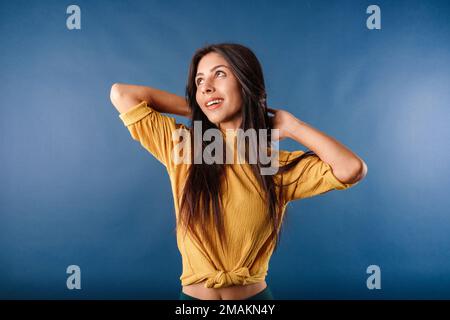 Porträt einer süßen, dunkelhaarigen Frau, die ein legeres Oberteil auf blauem Hintergrund trägt, entspannt und streckt, Arme und Hände hinter Kopf und Hals. Succ Stockfoto