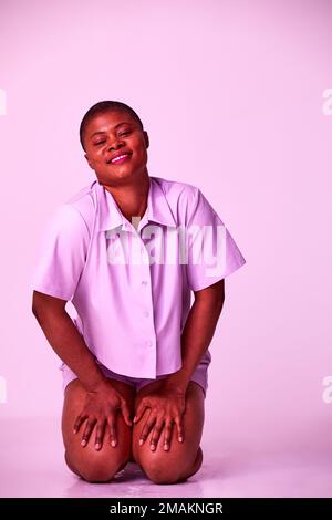 Eine übergroße, schwarze Millenial-Frau in lila T-Shirt und Shorts, die auf dem Boden sitzt und sich auf lila Studiohintergrund entspannen kann. Stockfoto