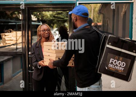 Pizzalieferung ins Büro, lächelnde Kunden, die Kartons stapeln, Bestellungen vom Kurier im Freien erhalten. afroamerikanischer Angestellter nimmt Essenspakete auf, Mann liefert Mittagessen Stockfoto