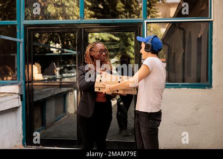 afroamerikanischer Kunde erhält Pizzabestellung im Freien, lächelnde Frau nimmt Essenspakete vom Kurier entgegen. Ein Mädchen mit Kopfhörern, das Essen zum Mitnehmen bringt, Pizzeria-Lieferservice Stockfoto