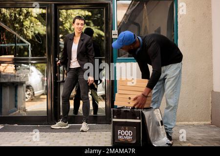 Der Kurier holt Pizzakartons aus dem Thermal-Rucksack heraus, der Kunde wartet draußen auf die Bestellung. Ein afroamerikanischer Mann, der Essen zum Mitnehmen bringt, ein Mitarbeiter des Unternehmens, der Fastfood bekommt Stockfoto