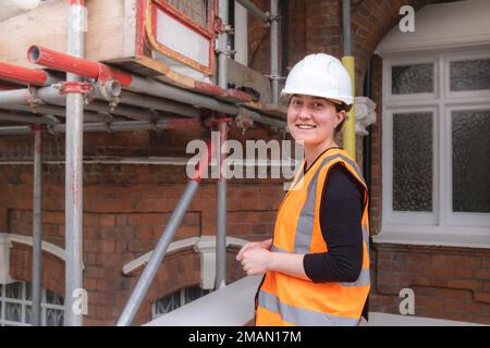 Eine lächelnde Bauingenieurin, die eine Inspektion durchführt und ein Baustellengebäude mit Gerüsten überwacht, einen Schutzhelm und Orange trägt Stockfoto