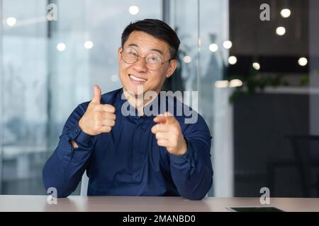 Ein erfolgreicher asiatischer Mann im Büro schaut auf die Webkamera, spricht mit Kollegen in einem Videogespräch, spricht und lächelt bei einem Online-Meeting mit Partnern, spricht mit Kunden, pov Stockfoto