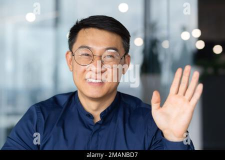Ein erfolgreicher asiatischer Mann im Büro schaut auf die Webkamera, spricht mit Kollegen in einem Videogespräch, spricht und lächelt bei einem Online-Meeting mit Partnern, spricht mit Kunden, pov Stockfoto