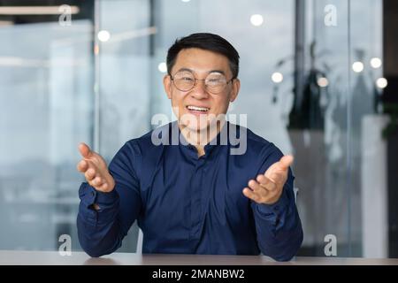 Ein erfolgreicher asiatischer Mann im Büro schaut auf die Webkamera, spricht mit Kollegen in einem Videogespräch, spricht und lächelt bei einem Online-Meeting mit Partnern, spricht mit Kunden, pov Stockfoto