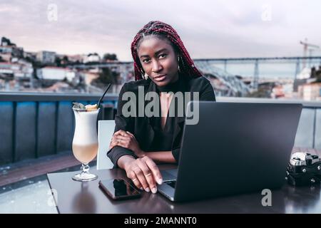 Eine faszinierende schwarze Universitätsstudentin in einer Bar auf dem Dach, die ihren Laptop benutzt, während sie an einem bewölkten Tag in Porto einen Cocktail auf einem unscharfen Blick genießt Stockfoto