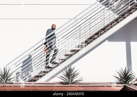 Ein schwarz bärtiger Geschäftsmann klettert unter dem Sonnenlicht die weiße metallische Treppe hinauf, trägt einen Anzug und hält eine Handtasche in einer minimalistischen Umgebung Stockfoto