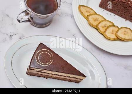 Ein Stück Schokoladenkuchen auf einem Teller mit einer Tasse Kaffee und Bananenscheiben auf dem Foto wird gemacht Stockfoto