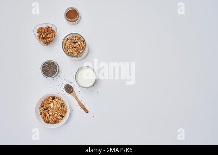 Einige Lebensmittel auf einer weißen Oberfläche mit Löffeln und Schüsseln gefüllt mit verschiedenen Sorten Müsli, Nüssen, Milch und anderen Zutaten Stockfoto