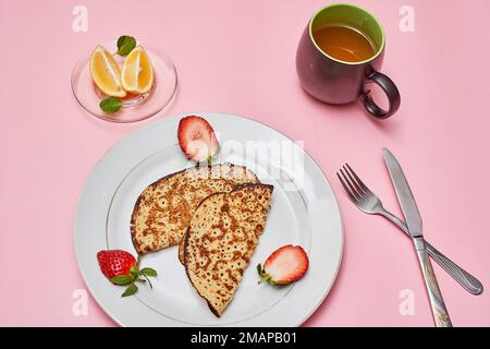 Zwei Pfannkuchen auf einem Teller mit Erdbeeren daneben und eine Tasse Tee im Hintergrund Stockfoto