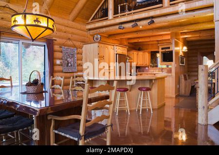 Esszimmer mit Holztisch und -Stühlen, Holzplankeninsel mit Keramikfliesen und Barhockern mit rotem Lederpolster in der Küche. Stockfoto