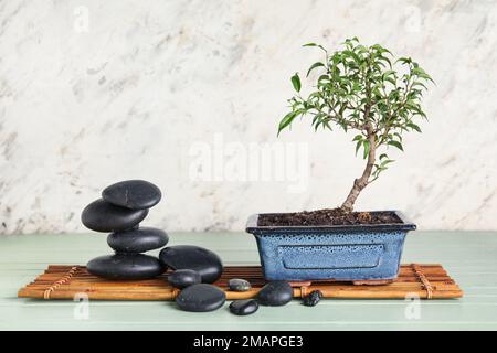 Bambusmatte mit Bonsai-Baum und Steinen auf dem Tisch nahe der hellen Wand Stockfoto