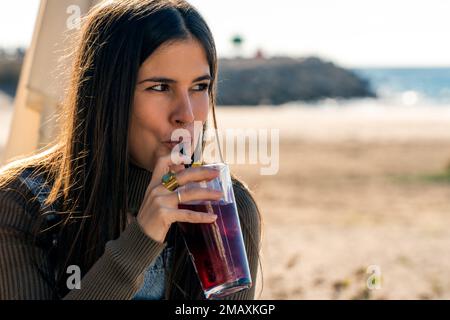 Junge Frau in legerer Kleidung trinkt frisches rotes kaltes Getränk aus Glas, während sie bei sonnigem Wetter Zeit an der Küste verbringt Stockfoto