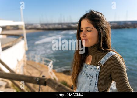 Hispanische Frau mit langen Haaren, die die Augen wunderbar schließt, während sie am Pier neben dem Meer steht und die Sonne genießt Stockfoto