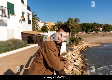 Junger Mann mit positivem Bart in brauner Jacke, der sich auf den Zaun lehnt, während er neben dem Meer und exotischen Bäumen unter wolkenlosem blauem Himmel steht und in die Kamera schaut Stockfoto