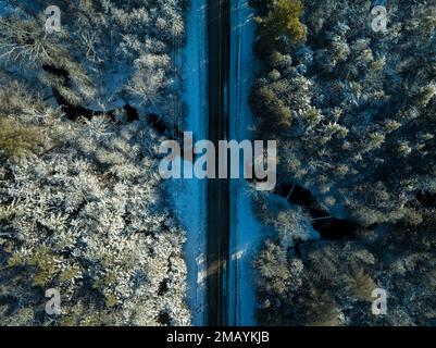 Ein Luftfoto über einer zweispurigen, geraden Straße, die über einen Bach in einer ländlichen Gegend durch einen gefrorenen, schneebedeckten Wald führt. Stockfoto