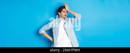 Verärgert Mann macht Facepalm und schaut besorgt weg, vergessen etwas wichtiges, schlagen Stirn beunruhigt, stehen vor blauem Hintergrund. Stockfoto