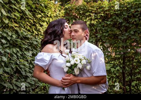 Ein Mädchen in weißem Kleid umarmt sanft den Hals ihres Freundes und küsst ihn auf die Wange. Frisch Vermählte im Park. Porträt Stockfoto