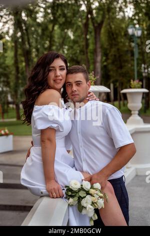 Ein Mädchen in hellweißem Kleid mit Blumenstrauß sitzt im Sommerpark auf dem Treppengeländer und umarmt ihren jungen Mann im weißen Hemd. Stockfoto