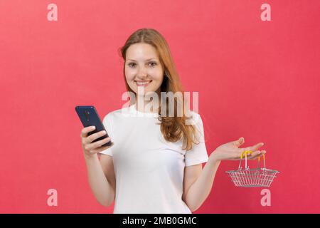 Wunderschöne junge weiße Frau, die einen kleinen Einkaufswagen und ein Mobiltelefon in einem weißen T-Shirt hält, um ein Modell zu erstellen, isoliert auf pinkfarbenem Hintergrund, Online-Shopping-Konzept Stockfoto