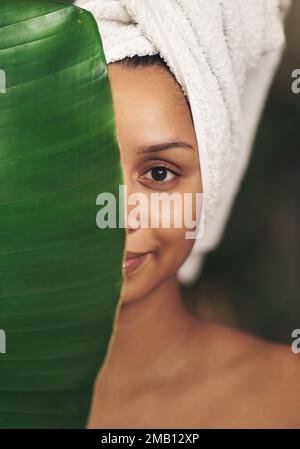 Ihre Haut bevorzugt natürliche Inhaltsstoffe. Eine schöne junge Frau, die ein Handtuch um den Kopf trägt, während sie hinter einem grünen Blatt posiert. Stockfoto