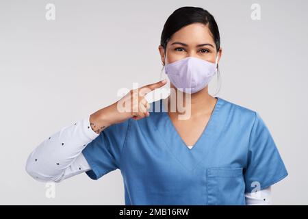 Nicht, was ich tue, sondern wer ich bin. Porträt eines jungen Arztes, der auf ihre chirurgische Gesichtsmaske zeigt, vor weißem Hintergrund. Stockfoto