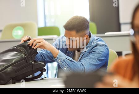 Universitätsstudent, Mann und Rucksack in einem Klassenzimmer für Bildung, Studium und Lernen auf dem Campus. Person in der Klasse, die nach Objekt im Beutel bis sucht Stockfoto
