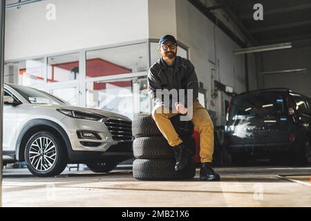 Ein Mann sitzt auf einem Stapel Autoräder in einer modernen und gut ausgestatteten Autoreparaturwerkstatt. Hochwertige Fotos Stockfoto