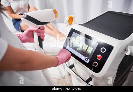 Nahaufnahme einer Kosmetikerin in sterilen Handschuhen mit einem Diodenlaser-Haarentfernungsgerät, während eine Frau am Tagesbett sitzt. Asthetiker-Vorrichtung für das Epilierverfahren. Stockfoto