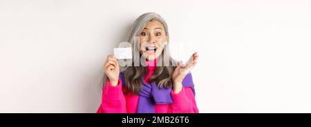 Einkaufskonzept. Glücklich asiatische alte aldy suchen beeindruckt und zeigt Kunststoff-Kreditkarte ihrer Bank, über weißem Hintergrund stehen Stockfoto