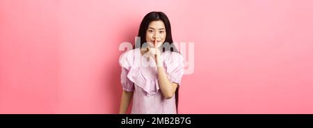 Nettes asiatisches Mädchen versteckt Geheimnis, mit Finger auf die Lippen gedrückt und lächelnd, bitten, ruhig zu bleiben, im Kleid auf rosa Hintergrund stehen Stockfoto