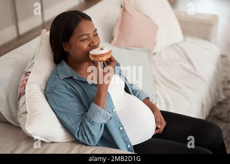 Sich ihrem Schwangerschaftsverlangen hingeben. Eine schwangere Frau, die zu Hause einen Donut isst. Stockfoto