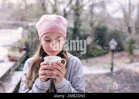 Wunderschönes Winterfoto eines Kindes, eines jungen Mädchens, eines Kindes im Pullover, das eine Tasse warmen Tee in der Hand hält. Mit Mütze fällt Schnee. Gemütlich, sieht friedlich aus Stockfoto