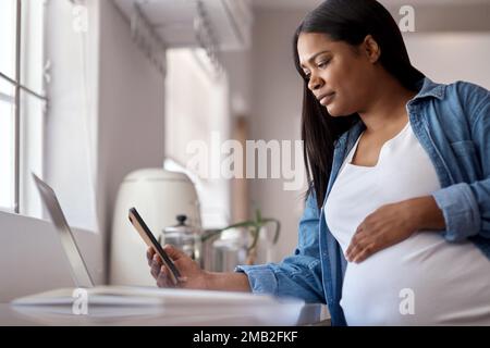 Ich habe noch ein paar Dinge zu erledigen, bevor mein Baby hier ist. Eine schwangere Frau, die ihr Handy benutzt, während sie zu Hause mit ihrem Laptop sitzt. Stockfoto