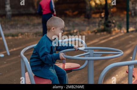 Süßer kleiner Junge mit Down-Syndrom in einem lustigen Hut läuft auf dem Spielplatz und dreht sich auf einem Karussell Stockfoto