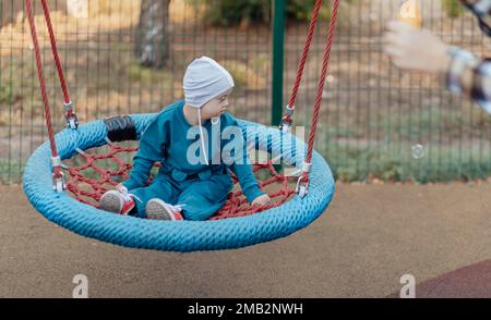 Süßer kleiner Junge mit Down-Syndrom in einem lustigen Hut läuft auf dem Spielplatz, schaukelt auf einer Schaukel Stockfoto