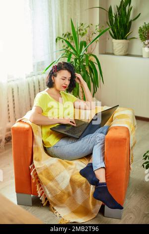 Frau, die sie benutzt, sucht ein Laptop, kauft online, bestellt Lieferung, auf einem orangefarbenen Sessel zu Hause, überprüft soziale Medien, Konzept des Heimgartens, grüne Hure Stockfoto